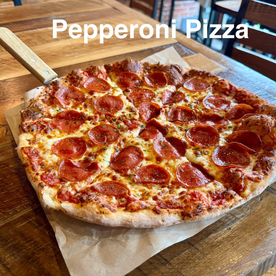 Pepperoni pizza with melted cheese and a crisp, charred crust sits on parchment atop a wooden restaurant table.