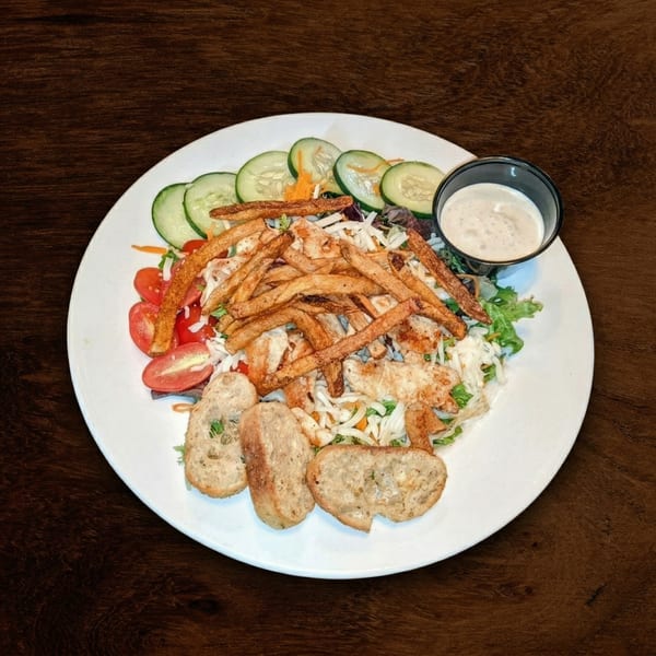 A white plate with a salad topped with chicken, fries, cucumbers, tomatoes, and garlic bread.