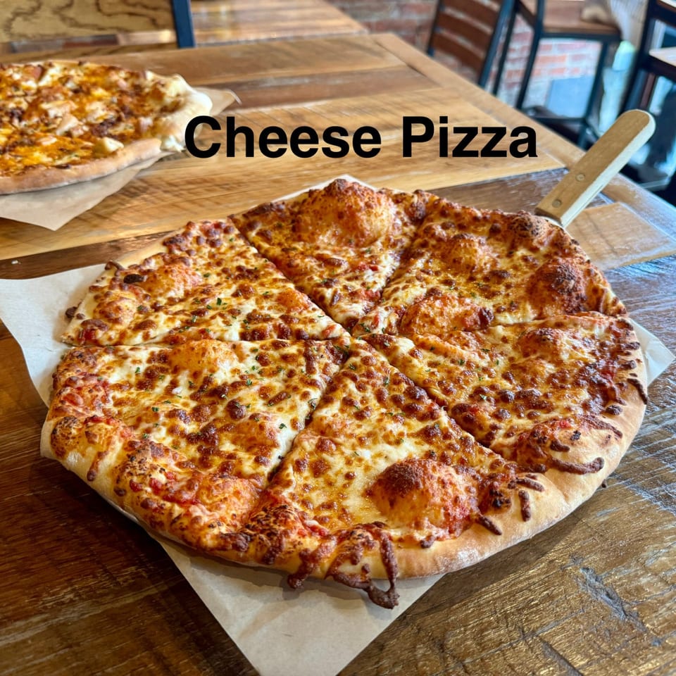 Large cheese pizza cut into slices on parchment paper atop a wooden restaurant table, with another pizza in the background.