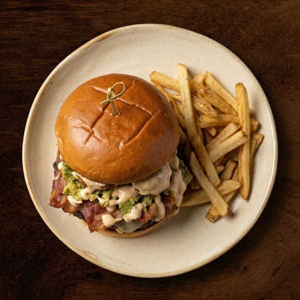 Gourmet burger with bacon, guacamole, and sauce, served with golden french fries on a white plate.