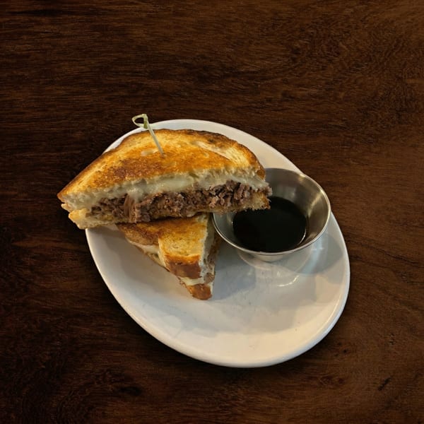 A grilled cheese sandwich with shredded meat and melted cheese, served with a side of au jus.