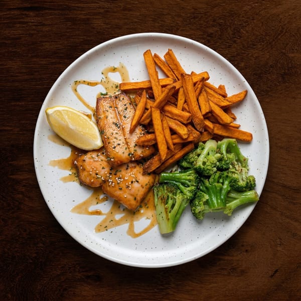 Salmon fillets with sesame seeds, sweet potato fries, broccoli, and a lemon wedge on a white plate.